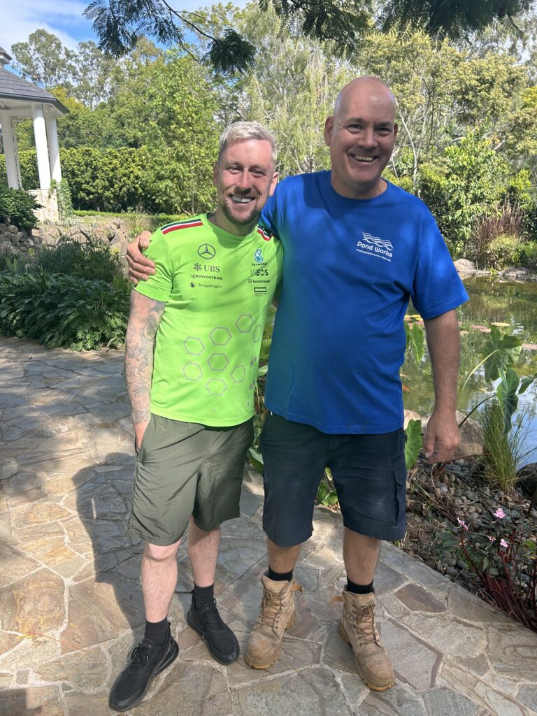 Mark Marshall visiting a Pond Works water feature project in Australia, standing beside a landscaped pond and waterfall with the Pond Works team.