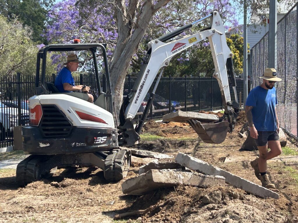 Excavator operated by Mark breaking up and removing concrete pathway at Kallangur State School during pond construction site preparation