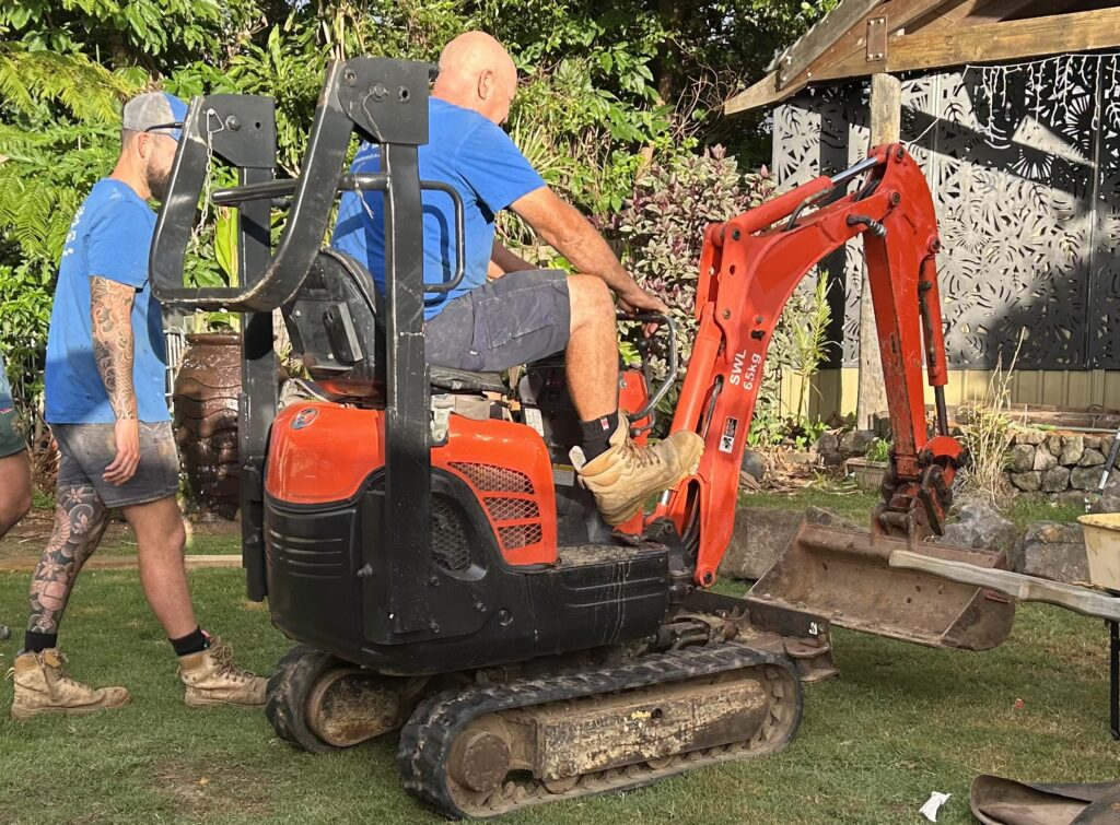 Two men in blue shirts watch as a bald operator drives a small orange excavator in a backyard yard.