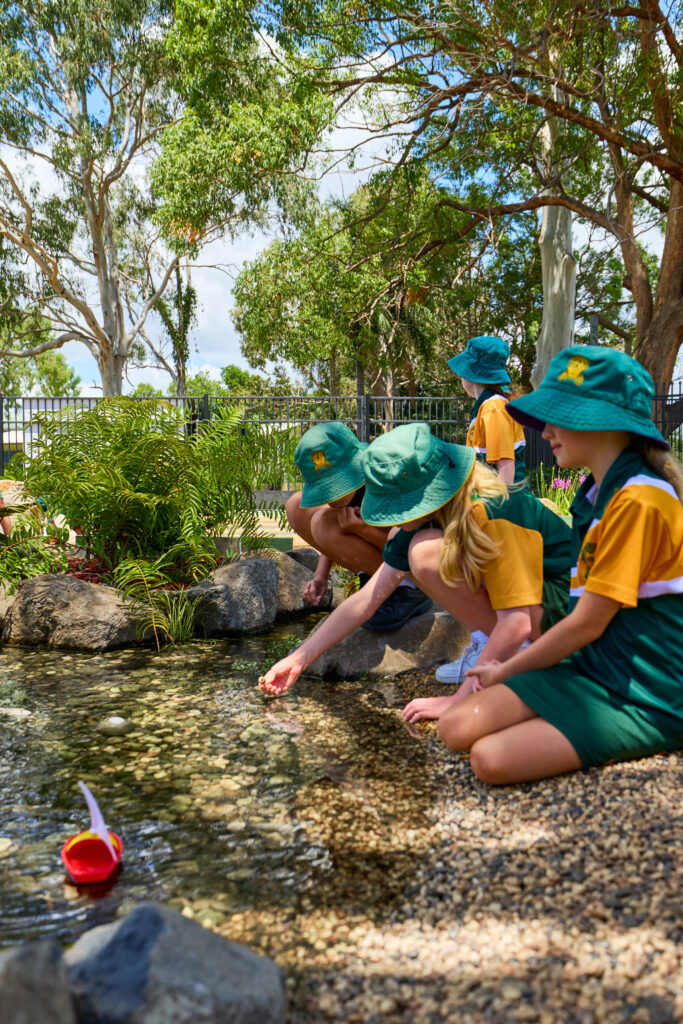 Group of children in green and yellow uniforms and teal hats kneeling by a pebbled pond, dipping hands into the water.