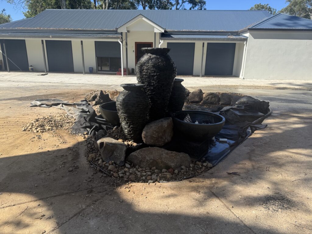 Construction of a custom ecosystem pond and decorative water feature in Thornlands, showing stacked slate water features being installed with the house in the background.